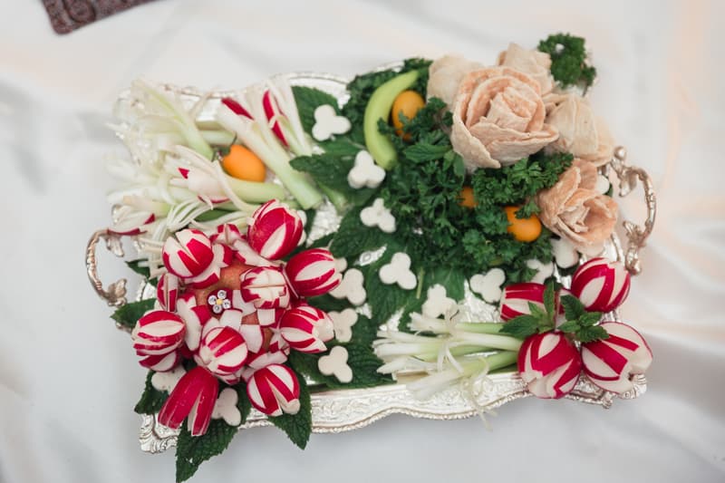 Silver tray with an arrangement of carved radishes, green herbs, and other vegetables on a white fabric surface.