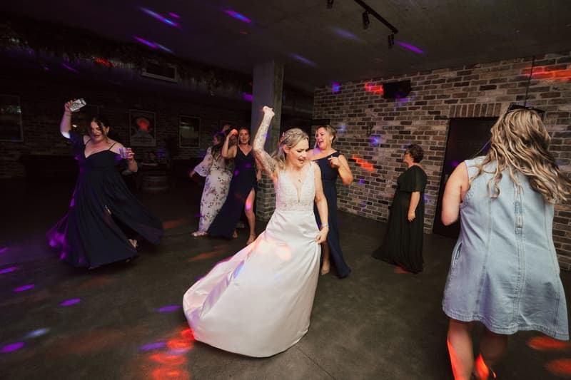 The bride Mandy dances energetically on the reception dance floor at Sandstone Point Hotel — The Cellar, surrounded by female guests and bridesmaids in dark dresses, with colorful disco lights illuminating the brick-walled room.