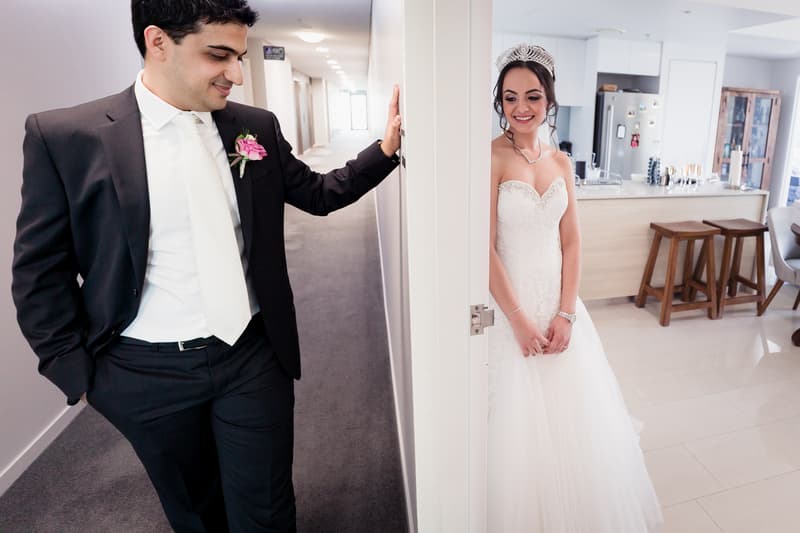 The groom, Pasha, in a black suit with a white tie and pink boutonniere, stands in a hallway leaning against a doorframe, while the bride, Maryam, in a strapless white wedding gown and tiara, stands on the other side of the doorframe in a kitchen area.