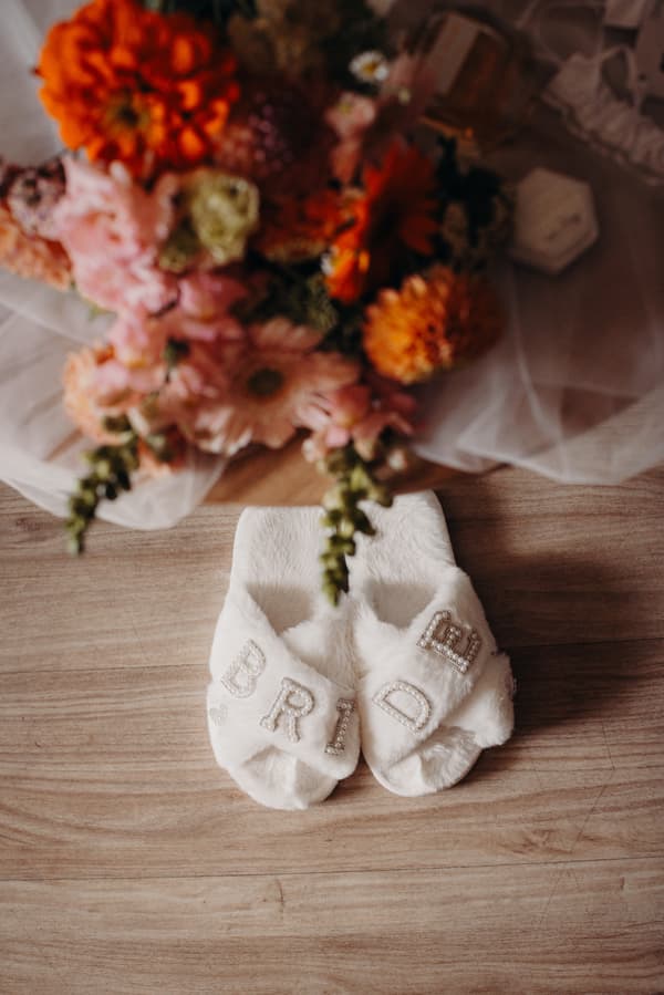 White fuzzy slippers with the word 'BRIDE' spelled out in pearl letters placed on a wooden floor next to a colorful bridal bouquet and a small hexagonal ring box at Sandstone Point Hotel.