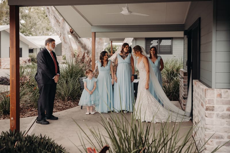 The bride Courtney stands with three bridesmaids and a flower girl in light blue dresses under a covered patio at Sandstone Point Hotel — Pavilion, while an older man in a suit and red striped tie watches.