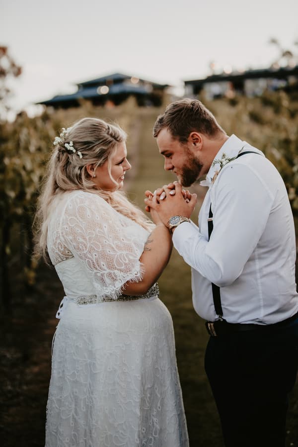 Bride Annie and groom Trent hold hands facing each other outdoors at Ocean View Estates, surrounded by vineyard rows.