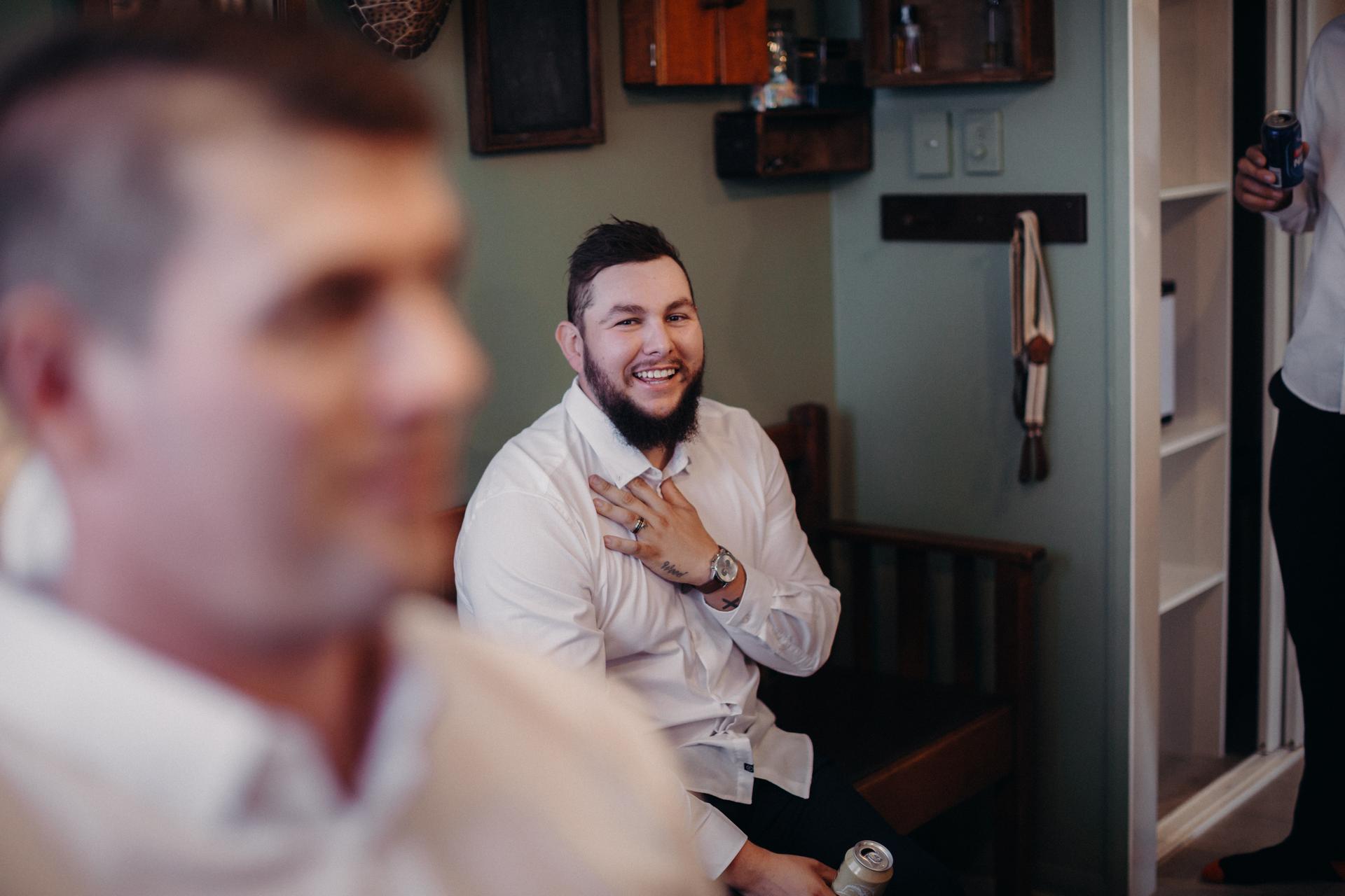 The groom Brent sits on a bench smiling and holding a can, with another man in the foreground out of focus, at Preston Peak Winery Functions Centre.