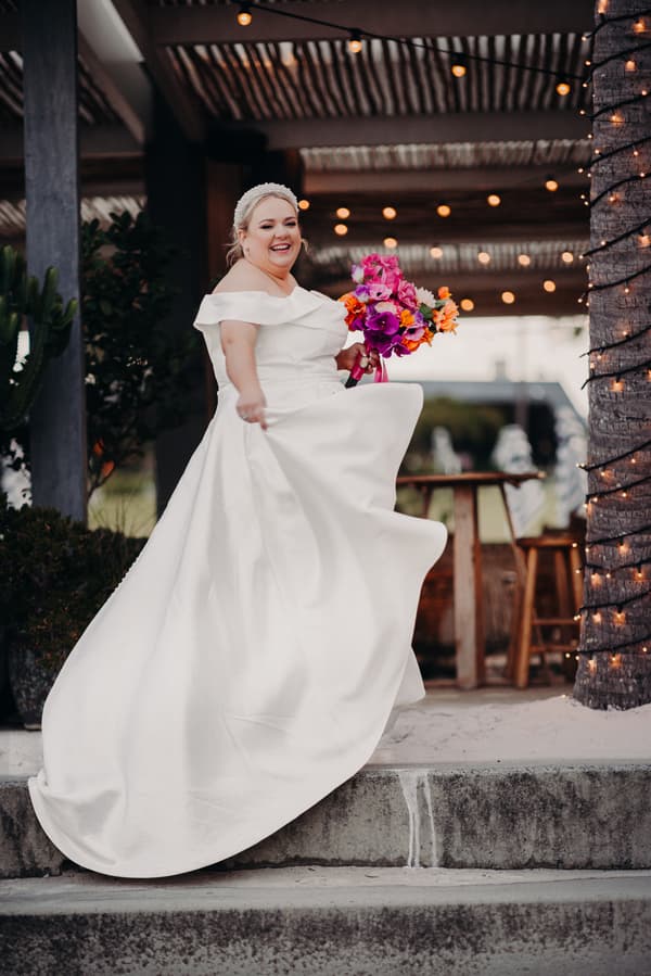 The bride Jacquelyne in a white off-shoulder wedding gown holds a vibrant bouquet of pink, purple, and orange flowers while standing on steps under a covered area at Sandstone Point Hotel.