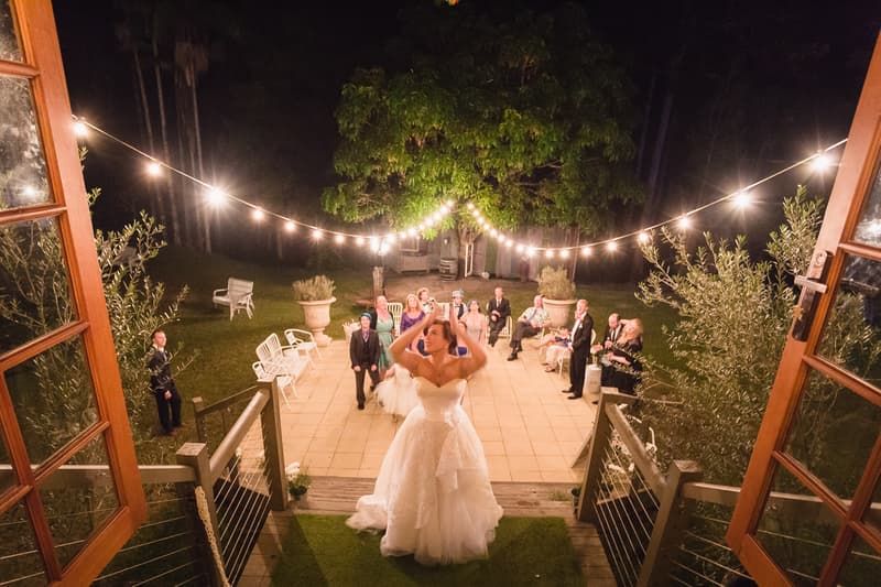 The bride Holly is tossing her bouquet to a group of guests standing on a patio at Kwila Lodge during the wedding reception.