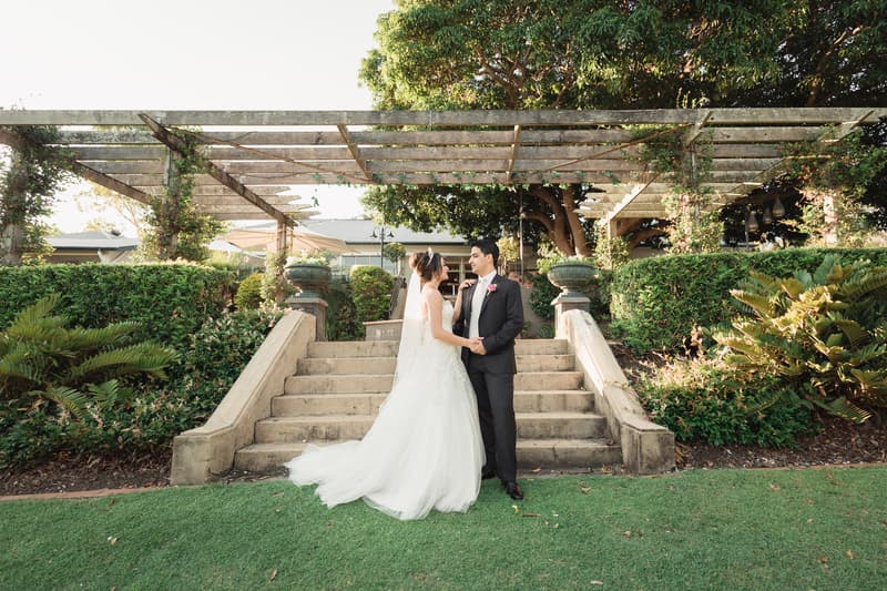 The bride and groom stand facing each other on the grass in front of stone steps and a pergola covered with greenery at Hillstone St Lucia — The Quartyard.