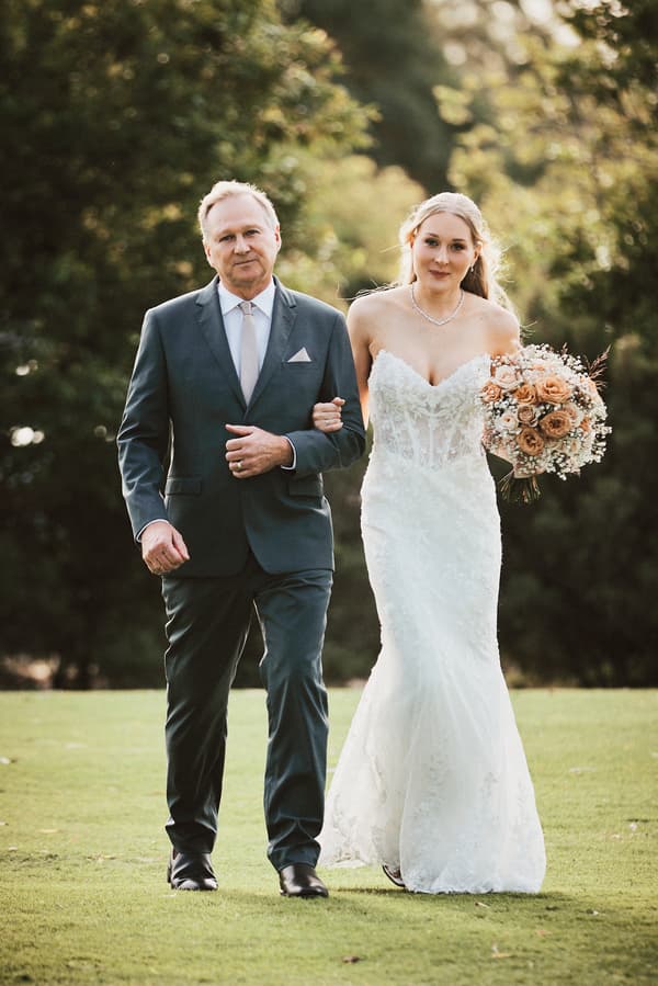 The bride Morgan, holding a bouquet of flowers, walks arm-in-arm with an older man, likely her father, on the lawn at Sandstone Point Hotel — The Pavilion.