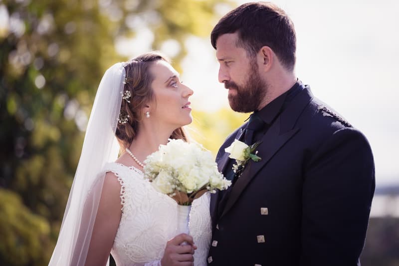 Bride Francesca and groom Ben stand close together outdoors, Francesca holding a white bouquet and wearing a veil and lace dress, Ben in a dark suit with a white boutonniere.