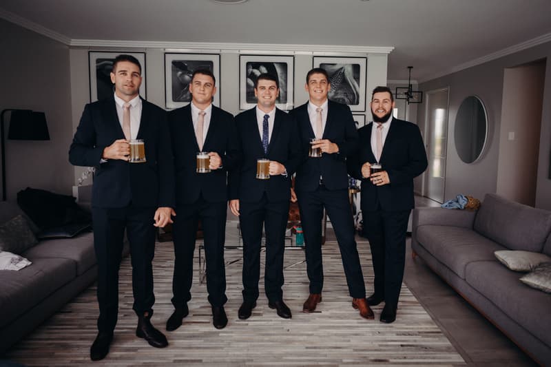 Five groomsmen stand indoors at Preston Peak Winery Functions Centre holding mugs of beer, dressed in dark suits with light pink ties except the man in the center who wears a dark blue tie with white dots.