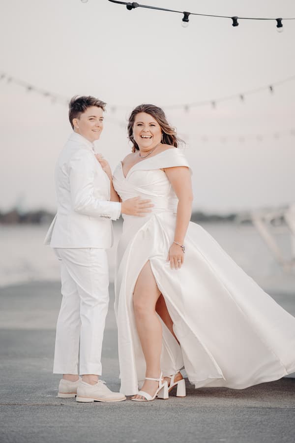 Brooke and Tiffany pose together outdoors, both dressed in white wedding attire, with string lights visible in the background.