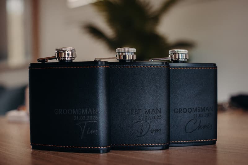 Three black leather flasks engraved with 'GROOMSMAN 21.02.2025 Tim', 'BEST MAN 21.02.2025 Dom', and 'GROOMSMAN 21.02.2025 Chris' placed on a wooden surface.