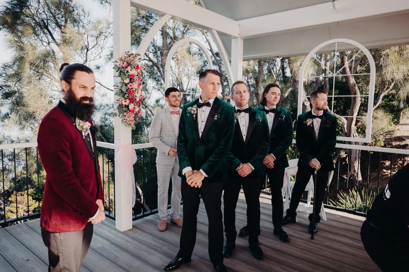 David and his groomsmen stand on the ceremony stage at Sandstone Point Hotel — Pavilion, dressed in tuxedos and suits, with floral decorations on the pillars behind them.