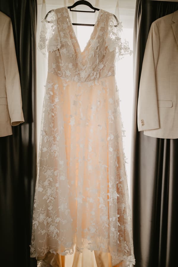 A wedding dress with floral lace detailing hangs on a black hanger in front of a window, flanked by two beige suit jackets hanging on either side.