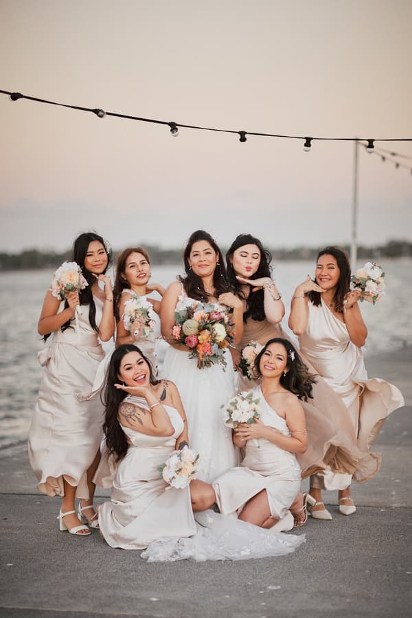 The bride Jheng poses with her bridesmaids in light-colored dresses holding bouquets near the water at Sandstone Point Hotel.