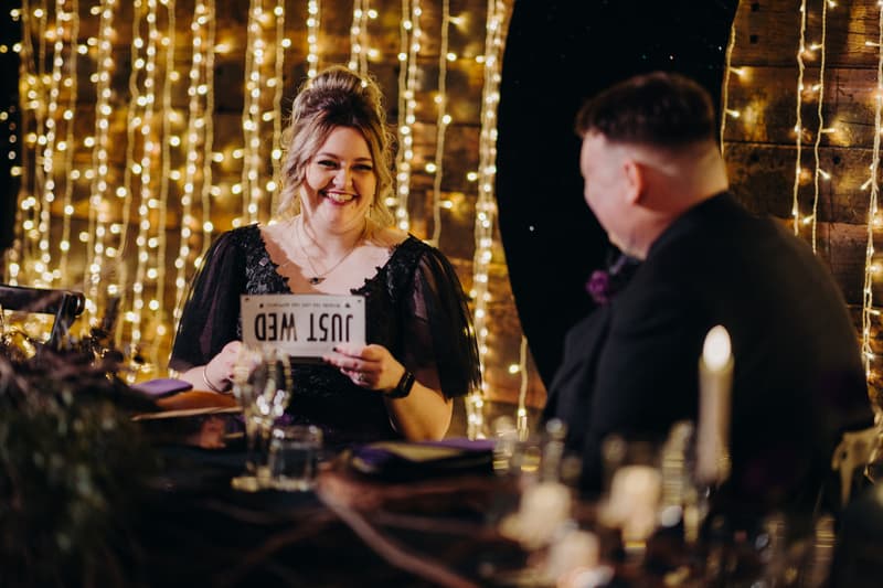The bride and groom sit at a table in the Ocean View Estates Function Room during the reception. The bride holds a 'JUST WED' sign and smiles, while the groom looks towards her. String lights decorate the wooden wall behind them.