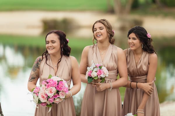 Three bridesmaids in matching champagne-colored dresses holding pink and white floral bouquets stand outdoors near a pond. Elopement photography Brisbane by She Said Yes.