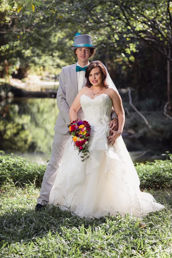 Bride Holly in a white wedding gown holding a colorful bouquet stands in front of groom Paul, who is wearing a light grey suit with a teal bow tie and a grey top hat with a teal band and feather, posing outdoors near a pond at Kwila Lodge.