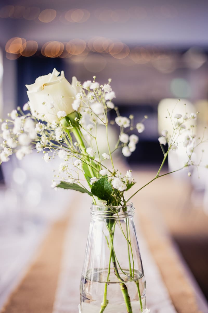 Close-up of a white rose and baby's breath flowers in a clear glass vase filled with water on a table at the. Getting ready photos by She Said Yes.
