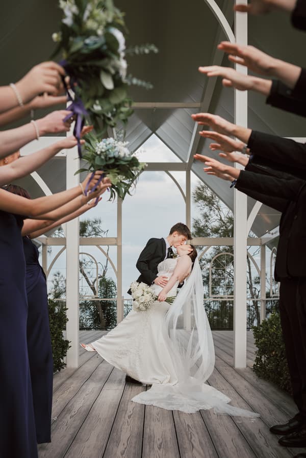 Tyler dips Terion for a kiss under the white pavilion structure at Sandstone Point Hotel — The Pavilion, surrounded by bridal party members holding bouquets and extending their arms.