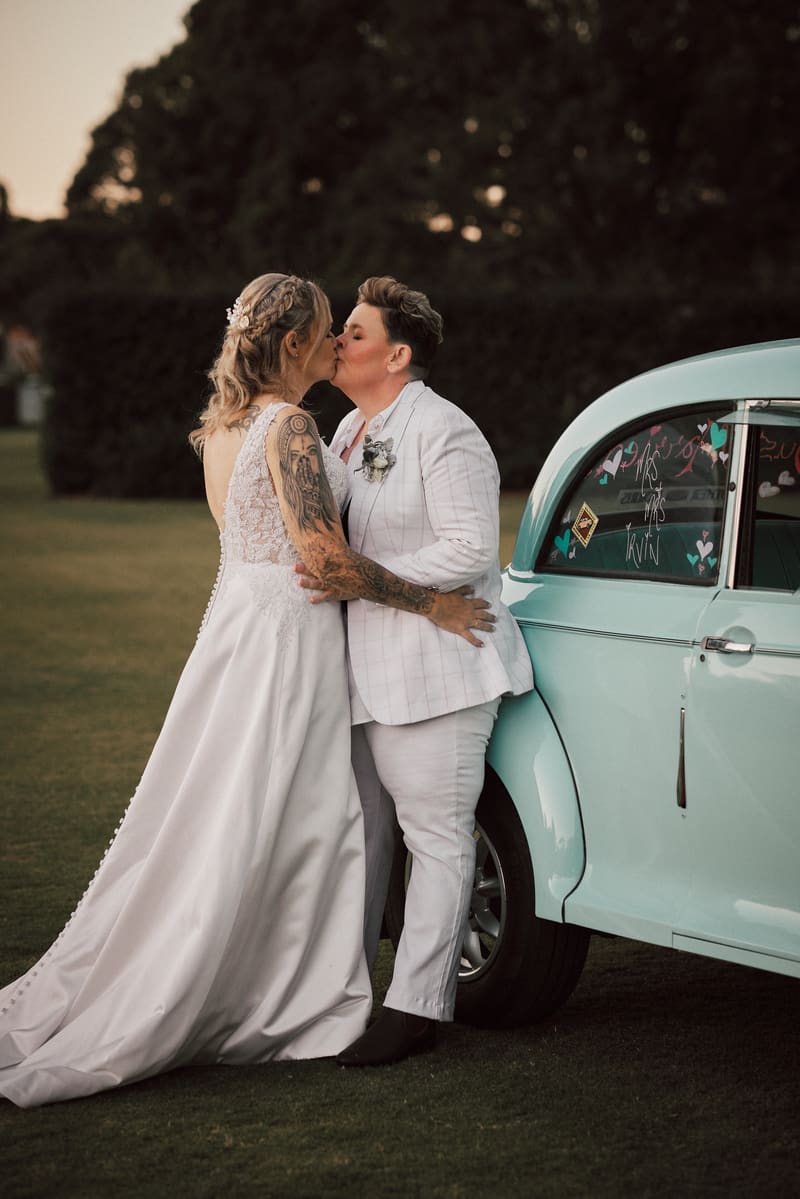 Mandy and Selina share a kiss outdoors beside a light blue vintage car. Full day wedding photography by She Said Yes.
