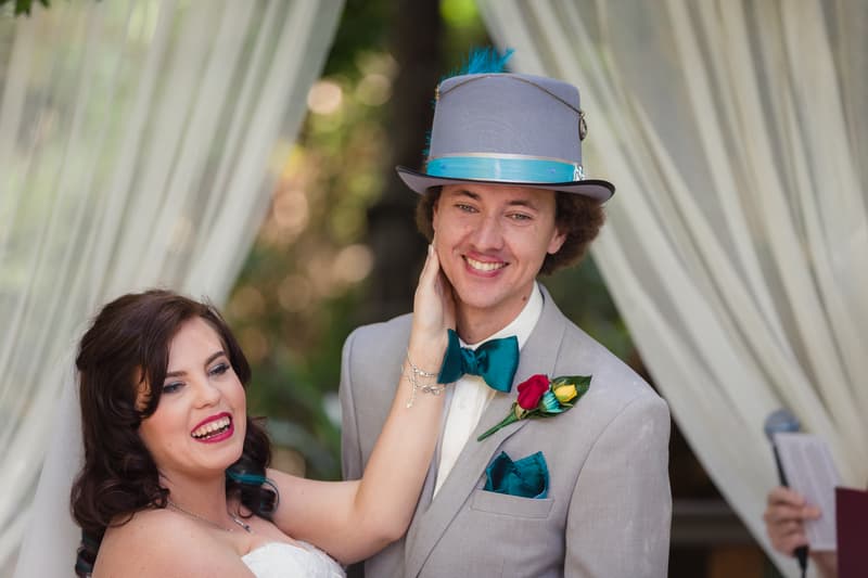 The bride Holly touches the groom Paul's face during their wedding ceremony at Kwila Lodge, with white draped fabric in the background and an officiant partially visible holding a book.
