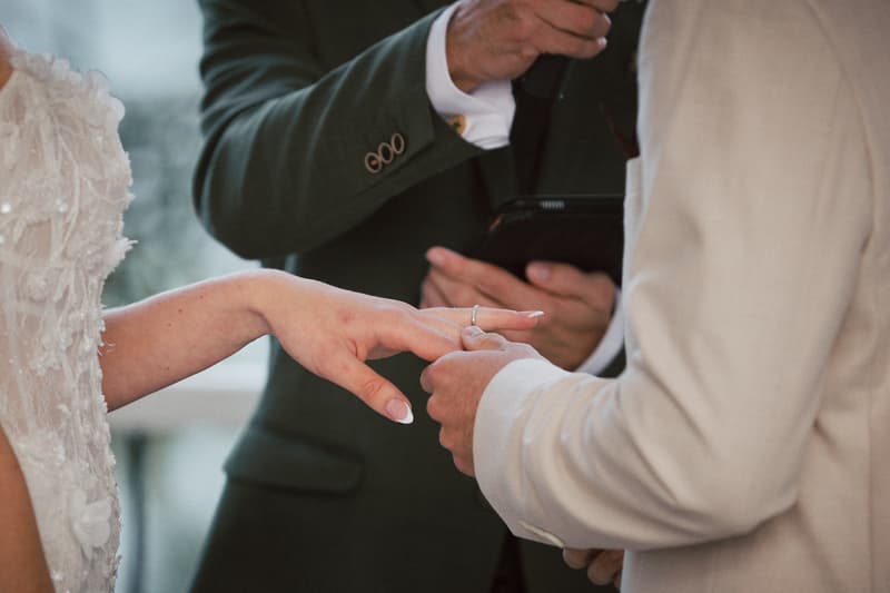 Joel places a wedding ring on Morgan's finger during the ceremony at Sandstone Point Hotel — The Pavilion, with the officiant holding a tablet in the background.