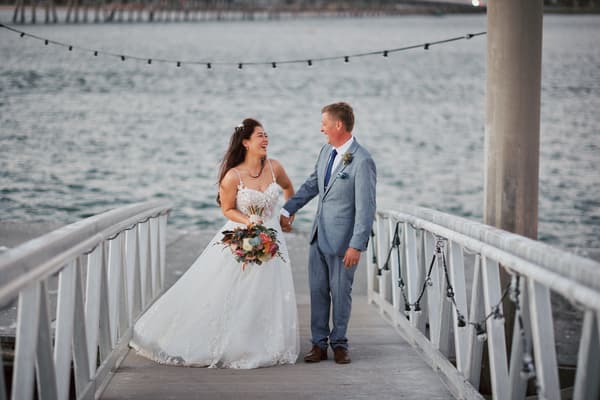 The bride Jheng in a white wedding gown holding a bouquet and the groom Dane in a light blue suit holding hands and smiling on a wooden pier with water in the background. ABIA Awards wedding photography by She Said Yes.