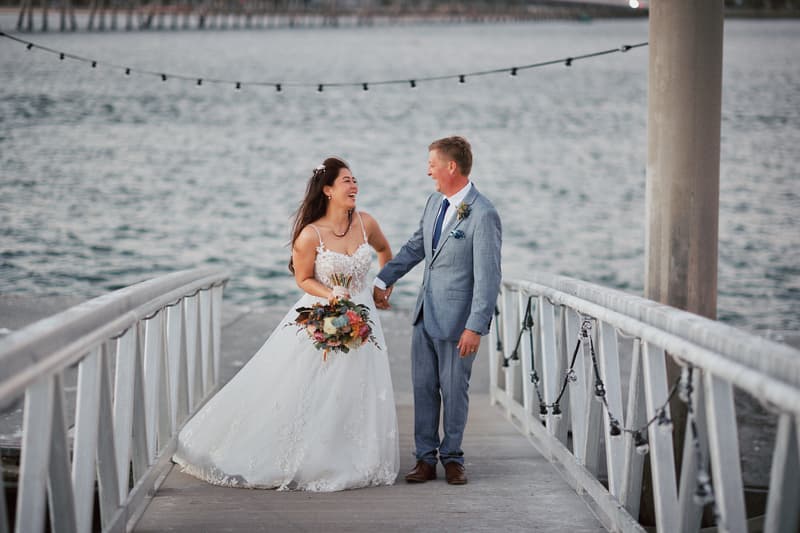 The bride Jheng in a white wedding gown holding a bouquet and the groom Dane in a light blue suit holding hands and smiling on a wooden pier at Sandstone Point Hotel with water in the background.