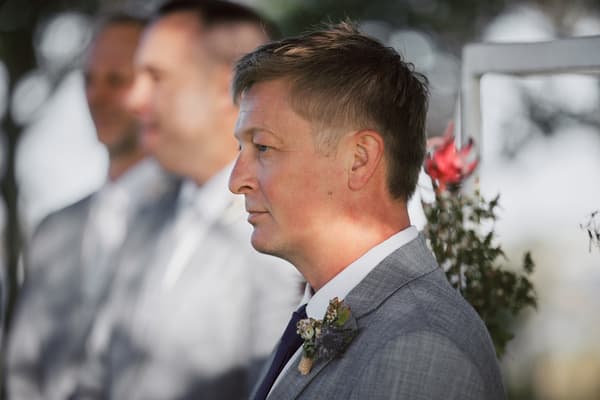 The groom stands in profile wearing a grey suit with a boutonniere, with groomsmen blurred in the background. Sandstone point hotel wedding by She Said Yes.