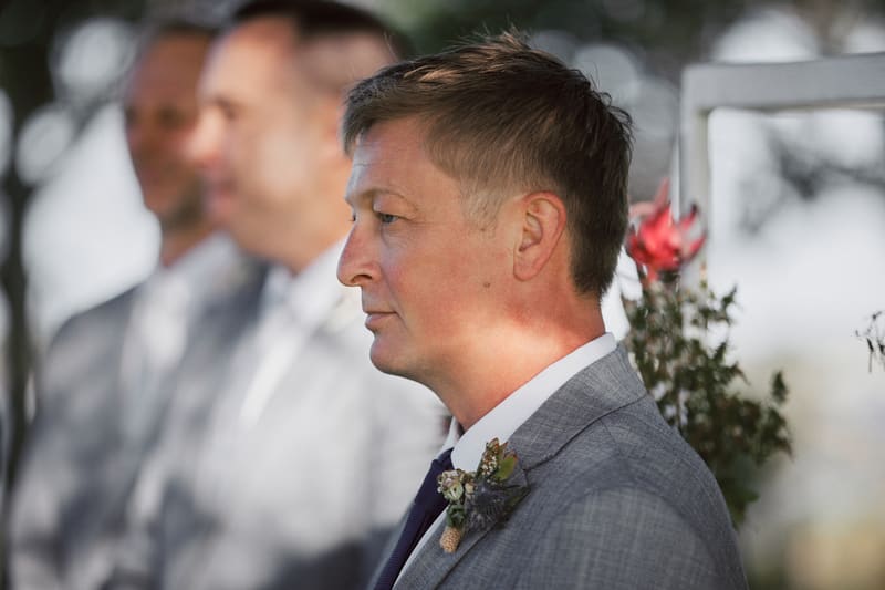 The groom stands in profile wearing a grey suit with a boutonniere, with groomsmen blurred in the background. Engagement photography Brisbane by She Said Yes.