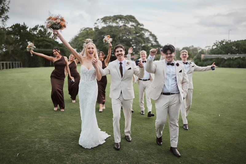 Morgan the bride and Joel the groom walk on grass at Sandstone Point Hotel with their bridal party, the bride holding a bouquet aloft and bridesmaids in brown dresses and groomsmen in cream suits following behind.