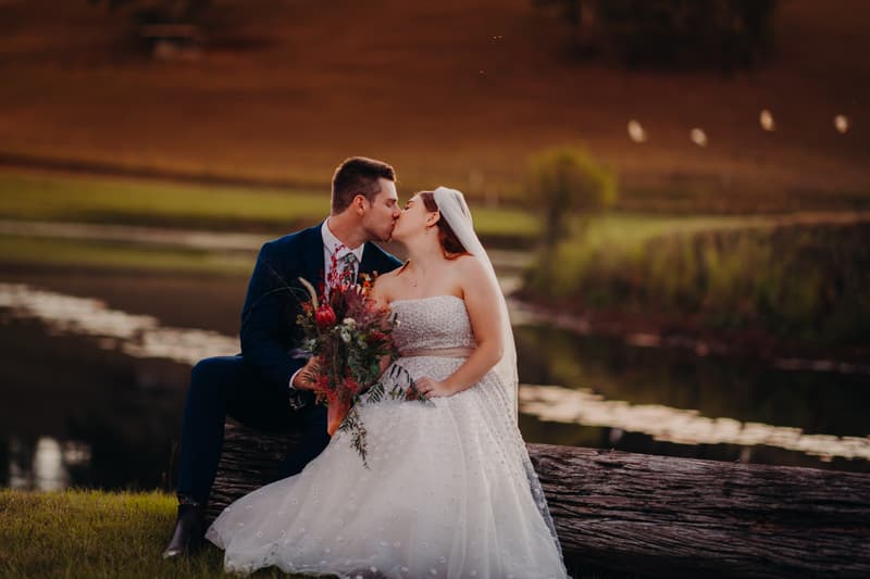 Bride Lilly and groom Connor sit on a log by a pond at Yabbaloumba Retreat, sharing a kiss. Lilly wears a strapless white wedding gown and veil, holding a bouquet, while Connor wears a dark suit.