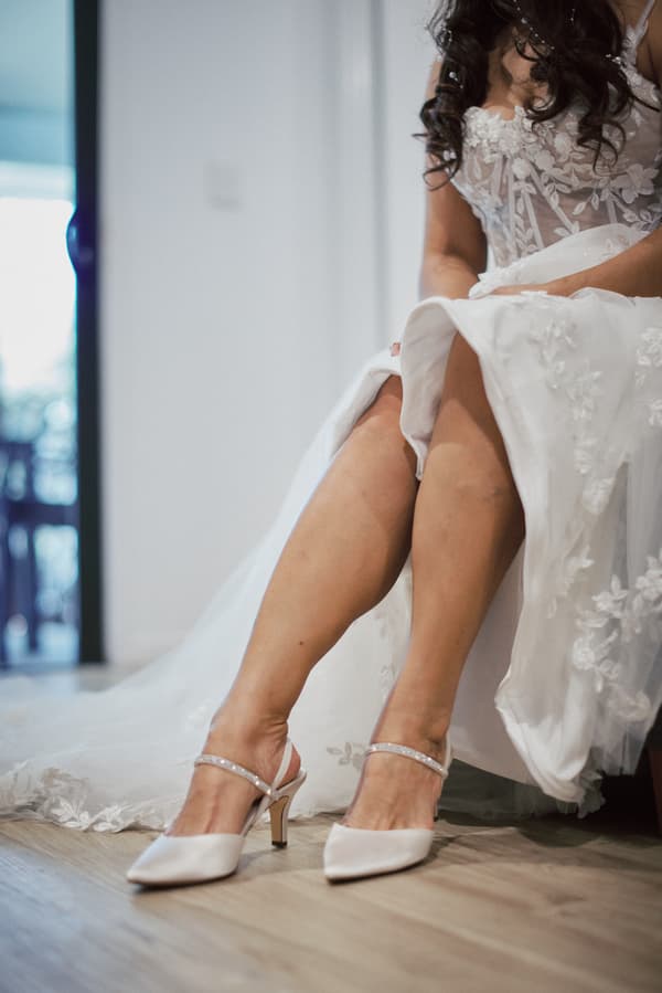 The bride sits indoors wearing a white lace wedding dress and white heeled shoes with ankle straps, showing her legs and part of the dress train.