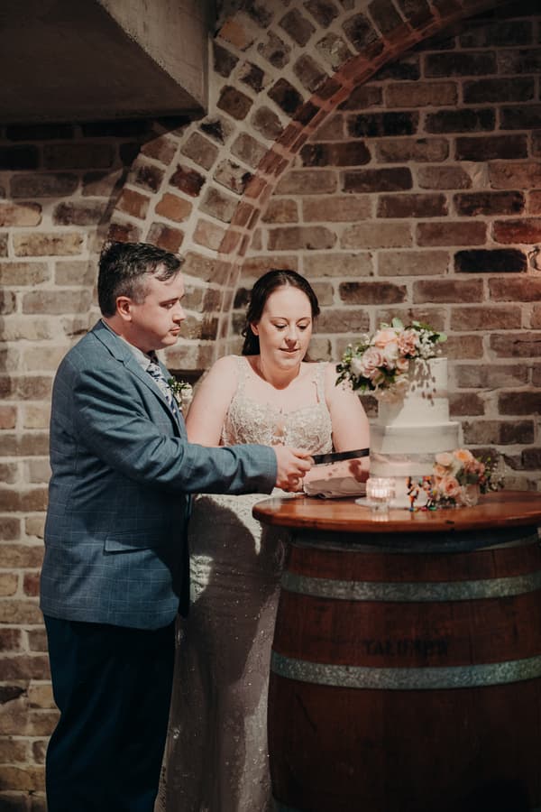 Rebecca and Dale cut their wedding cake on a wooden barrel table at the Sandstone Point Hotel — Cellar.