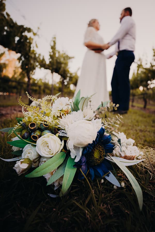 The bride and groom hold hands standing in a vineyard at Ocean View Estates, with a close-up of a bridal bouquet featuring white and blue flowers in the foreground on the grass.