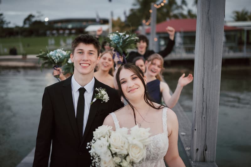 Tyler and Terion stand together on a wooden pier at Sandstone Point Hotelallo, smiling at the camera with the bridal party behind them holding bouquets and cheering.