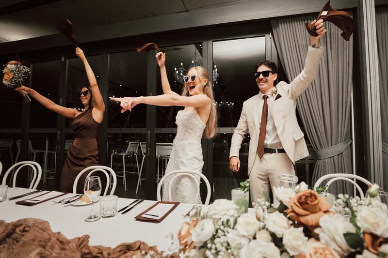 The bride Morgan in a white wedding dress, the groom Joel in a light suit, and a bridesmaid in a brown dress wave brown cloths and a bouquet at the reception in The Pumicestone Room at Sandstone Point Hotel.