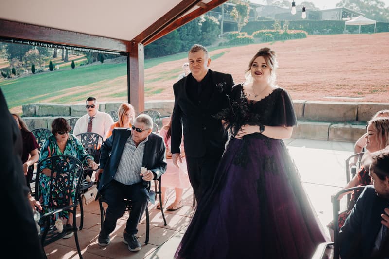 The bride Renae in a dark purple and black gown walks arm-in-arm with an older man, likely her father, down an aisle at Ocean View Estates — On The Lake, while seated guests watch.