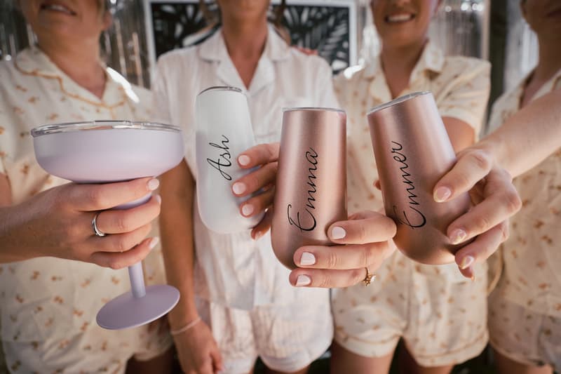 The bride Ashleigh and bridesmaids hold personalized drinkware with names Ash and Emma visible, wearing matching light-colored pajamas at Sandstone Point Hotel.