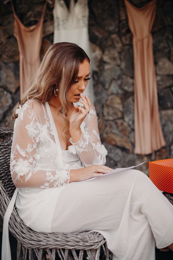 The bride Chelsea sits on a wicker chair wearing a white floral lace robe, reading a piece of paper with two bridesmaid dresses hanging on a stone wall behind her at Preston Peak Winery Functions Centre.