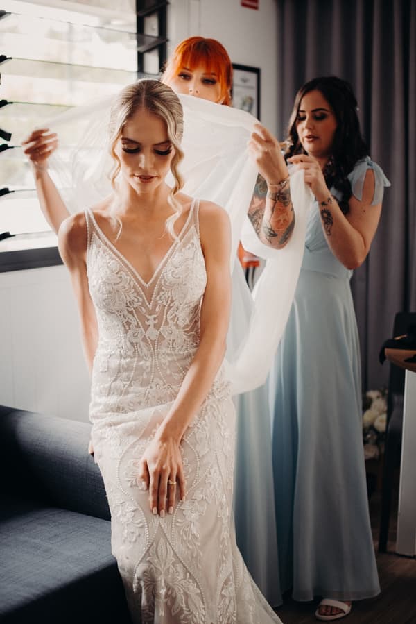 The bride Courtney is seated while two bridesmaids adjust her veil in a room at Sandstone Point Hotel.