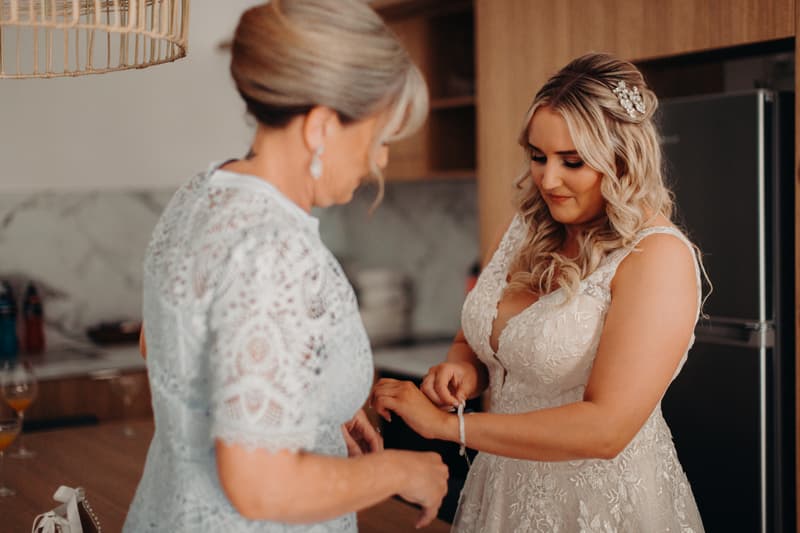 The bride Chloe is having a bracelet fastened on her wrist by an older woman, likely the mother of the bride, in a kitchen area at Sandstone Point Hotel.