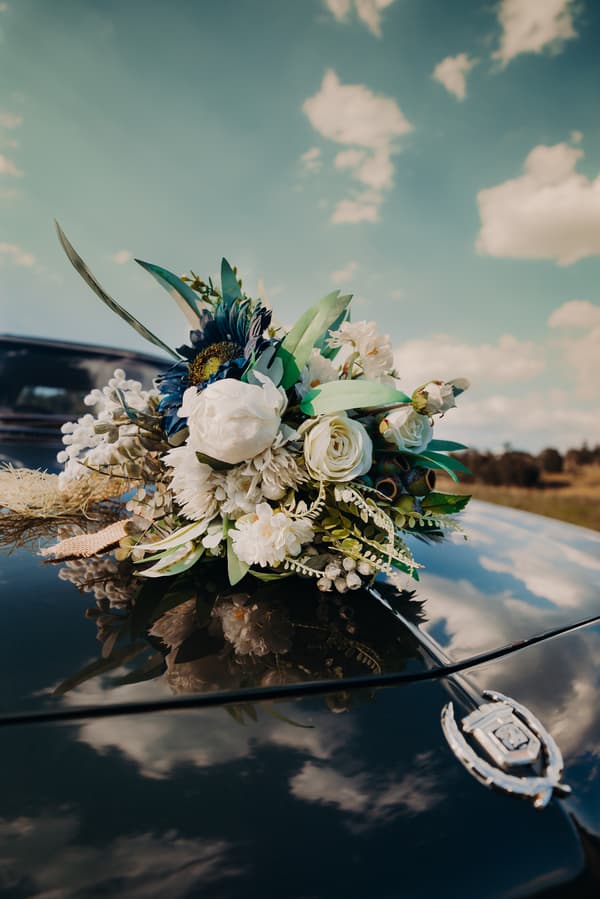 A bridal bouquet with white and blue flowers rests on the hood of a black vintage car at Ocean View Estates, reflecting the sky and clouds above.