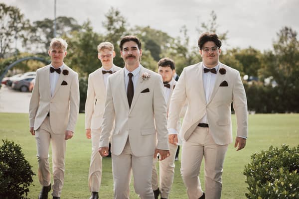Joel the groom walks outdoors on grass with four groomsmen, all wearing light beige suits with dark bow ties and boutonnieres. Shortest day wedding timeline by She Said Yes.