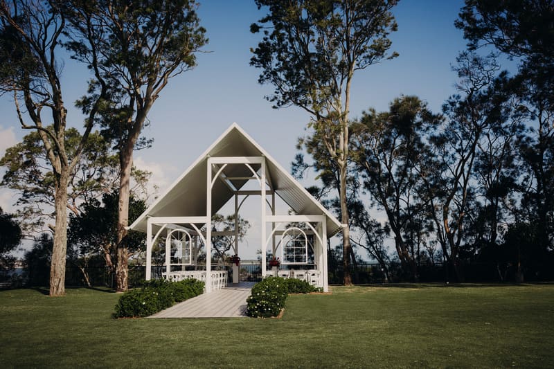 Empty white pavilion structure set up for a wedding ceremony at Sandstone Point Hotel with rows of white chairs and greenery on a lawn surrounded by tall trees under a clear sky.
