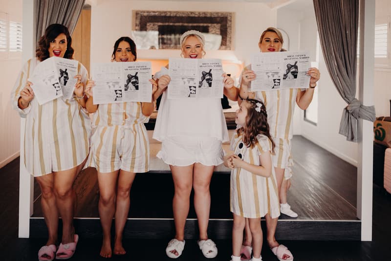 The bride Jacquelyne stands with three bridesmaids and a flower girl at Sandstone Point Hotel — Pavilion, all holding newspapers with the headline 'ARENA AND CO ARE GETTING MARRIED'.