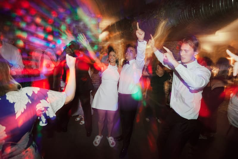 Guests, including a woman in a white dress and men in white shirts and bow ties, dance under colorful lights at the reception in Sandstone Point Hotel — The Cellar.