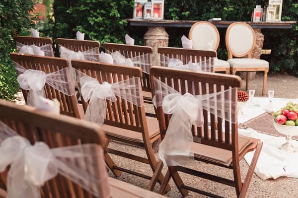 Rows of wooden chairs decorated with white tulle bows set up for a wedding ceremony at Hillstone St Lucia — The Quartyard, with a table and two chairs in the background and fruit bowls on a white cloth on the ground.