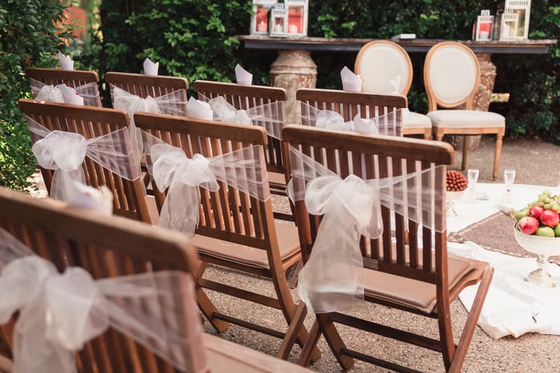 Rows of wooden chairs decorated with white tulle bows set up for a wedding ceremony at Hillstone St Lucia — The Quartyard, with a table and two chairs in the background and fruit bowls on a white cloth on the ground.