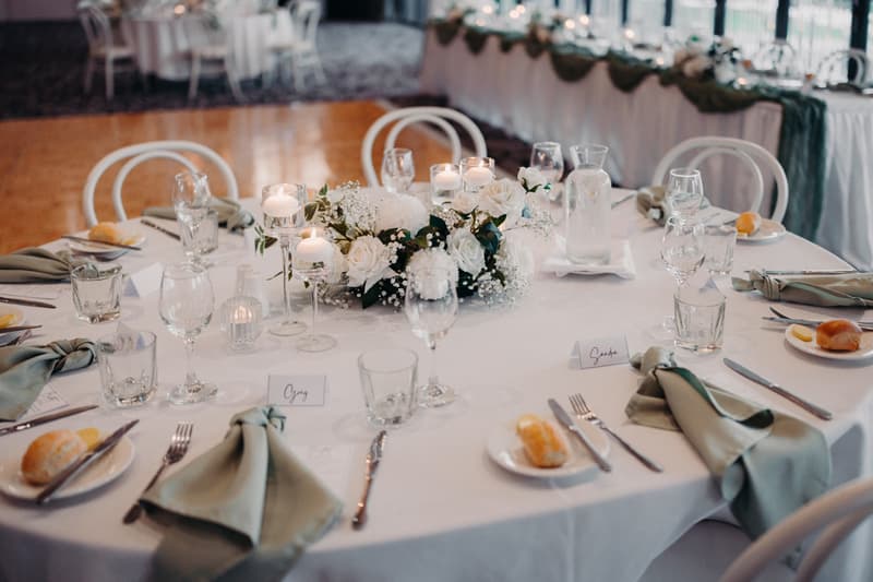 A round reception table at Sandstone Point Hotel — Pumicestone Room is set with white tablecloth, green napkins, glassware, bread rolls on plates, and a floral centerpiece with white flowers and candles.